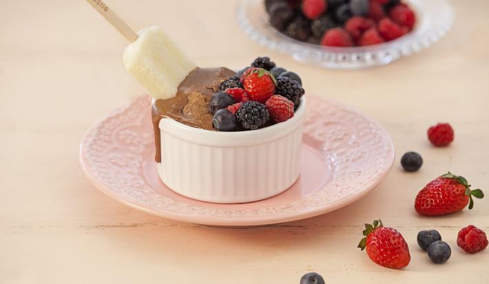 Fotografia em tons de rosa e branco de uma bancada rosa vista de frente, ao centro um prato redondo rosa e por cima um potinho branco com frutas vermelhas e um picolé mergulhando na calda de chocolate. Ao fundo um prato transparente com frutas vermelhas.