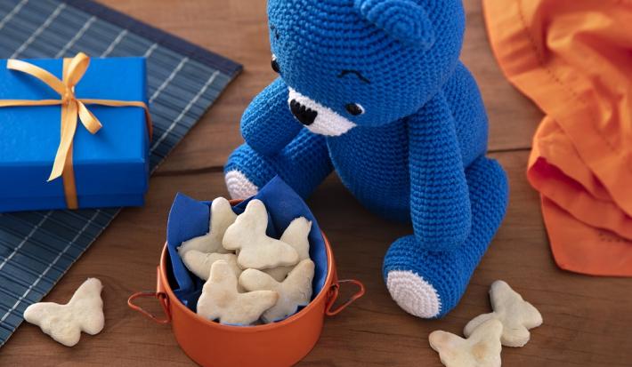 Fotografia em tons de branco em uma bancada de madeira de cor marrom. Ao centro, um recipiente laranja contendo os sequilhos. Ao lado, um urso azul, uma caixinha azul e ao fundo um pano laranja.