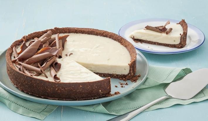 Fotografia em tons de azul em uma mesa de madeira azul com um prato raso azul claro e a torta de limão e chocolate. Ao lado, um prato pequeno com uma fatia da torta e uma espátula de bolo.