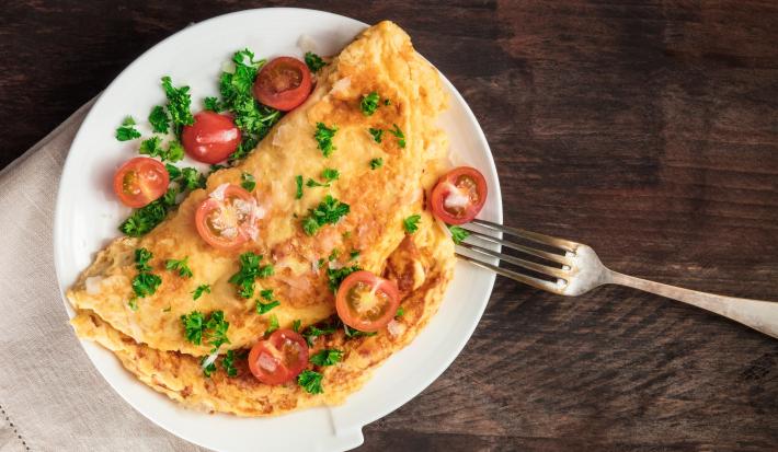 fotografia em tons de marrom, branco, amarelo e vermelho de uma bancada marrom vista de cima. Contém um prato redondo branco com uma omelete, e por cima pedaços de tomates e salsas