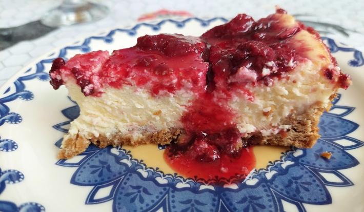 Foto em tons de vermelho da receita de cheesecake de frutas vermelhas ao estilo nova york servida em uma fatia sobre um prato decorado de porcelana azul e branco