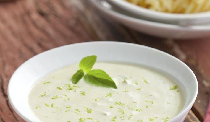 Fotografia com tons de branco e marrom, com ramequim branco com molho, ao fundo prato branco com macarrão parafuso e pedaço de queijo com ralador sobre tábua branca, ao lado guardanapo listrado, tudo sobre bancada de madeira rústica.,