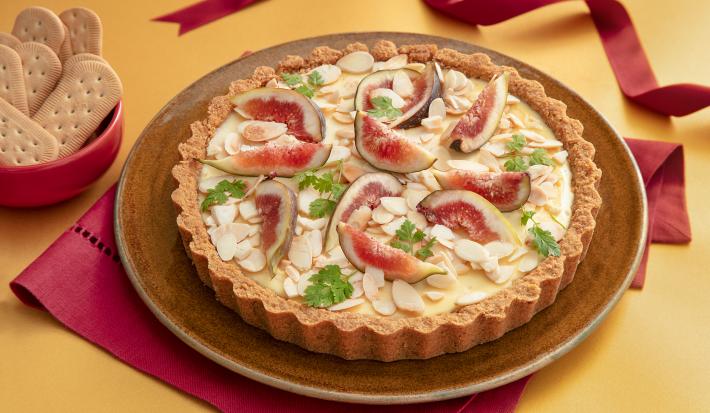 Foto da receita de  Torta de Figo, Ricota e Amêndoas. Observa-se a torta decorada com figos pela metade e amêndoas laminadas em uma cenografia amarela e vermelha. Ao lado esquerdo, biscoitos inteiros decoram a foto