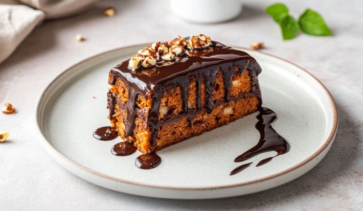 Fotografia de uma fatia de bolo com dourado e pedaços brancos, coberta com ganache de chocolate e nozes, servida em um prato cerâmico, com um toque de chocolate no fundo.