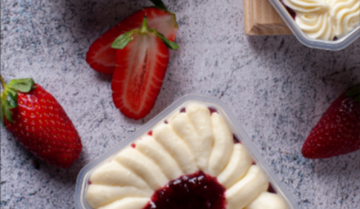 Imagem mostra, de cima, dois recipientes quadrados transparentes com um creme branco e um creme vermelho no topo. Estão dispostos em uma bancada de granito cinza, com uma tábua de madeira com morangos inteiros, cortados jogados.