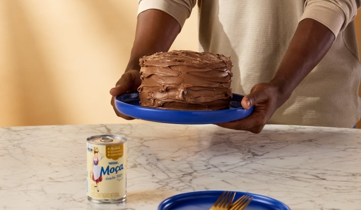 Fotografia mostra uma pessoa segurando um bolo com ganache de chocolate, com decoração rústica.