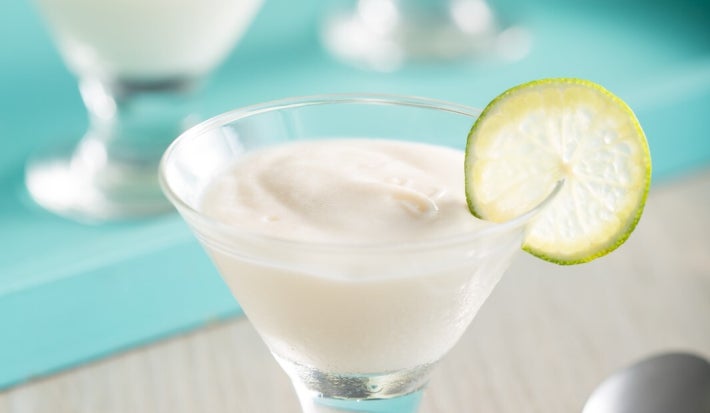 Foto de uma taça de vidro com sobremesa cremosa de limão, de cor clara e textura lisa. Uma fatia fina de limão está apoiada na borda. Ao fundo, há outras taças semelhantes sobre bandeja azul. A cena inclui colher prateada e mesa clara