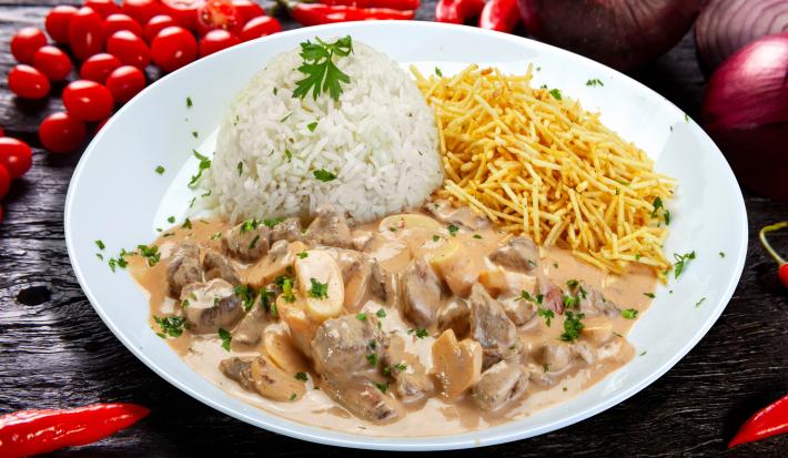 Imagem de um prato redondo branco com arroz, batata palha e strogonoff de carne cremoso. Ao fundo, cebolas e tomates cereja.