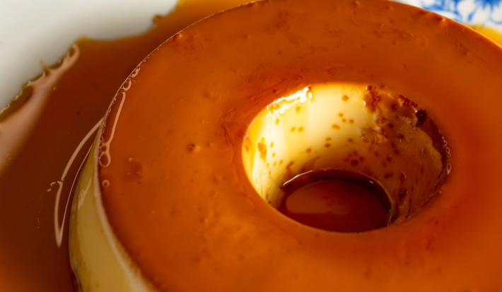 Fotografia de um prato branco, com desenhos azuis, grande raso sobre uma mesa de madeira. Sobre o prato tem um pudim pequeno tradicional feito com amido de milho na receita.