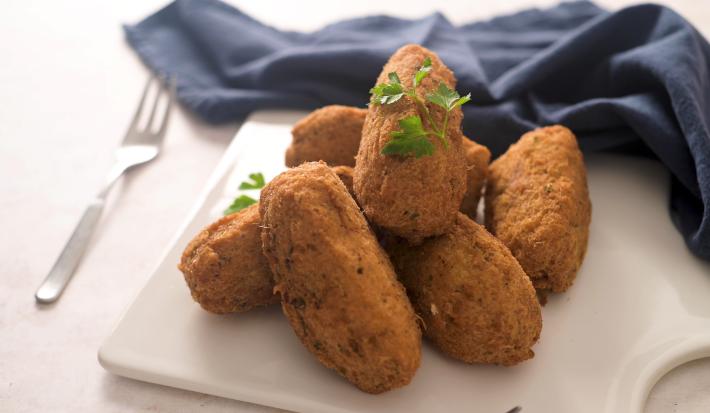 Fotografia de vários bolinhos de bacalhau fritos sobre um apoio branco, ao lado de um pano azul de tom escuro e um garfo.