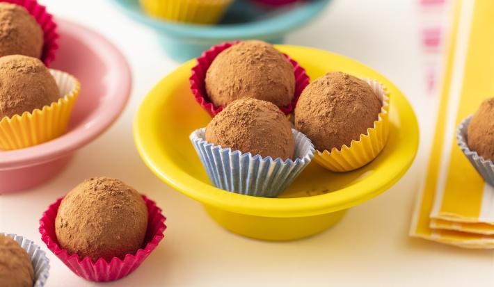 Fotografia em tons de marrom, em uma bancada de madeira branca. Ao centro, há um recipiente redondo amarelo contendo 3 bananas trufadas dentro de forminhas. Ao lado, há algumas bananas trufas espalhadas e um pano listrado amarelo.