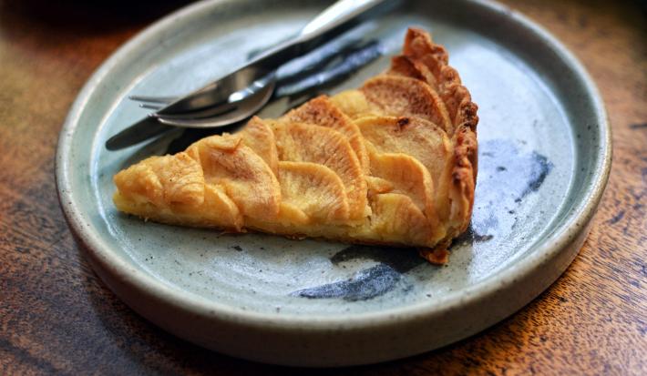Fotografia de uma fatia de torta de maçã caramelada e recheada com pedaços de maçã. Ao lado da torta tem talheres que também estão por cima de um prato raso de cerâmica em tom de azul, e ele está sobre uma mesa de madeira.