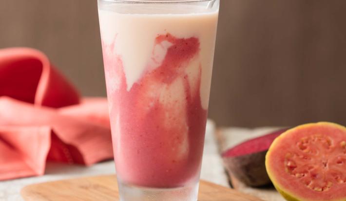Fotografia em tons de rosa em uma bancada de madeira, um pano rosa, uma tábua de madeira, um copo de vidro alto com a bebida de iogurte com goiaba, morango e beterraba. Ao lado, uma goiaba cortada ao meio.
