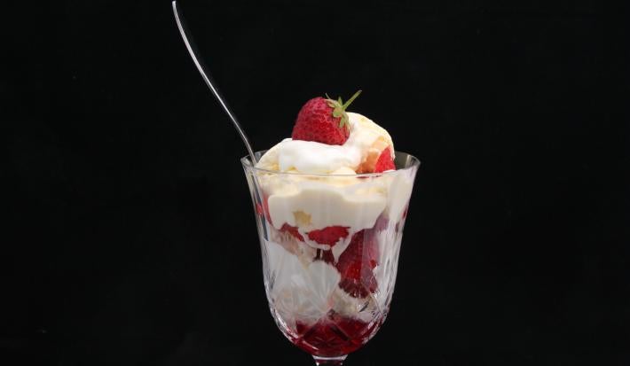 Fotografia de uma taça de vidro com merengue e morangos, de forma intercalada. E dentro do recipiente, uma colher de sobremesa apoiada, sobre um fundo preto.