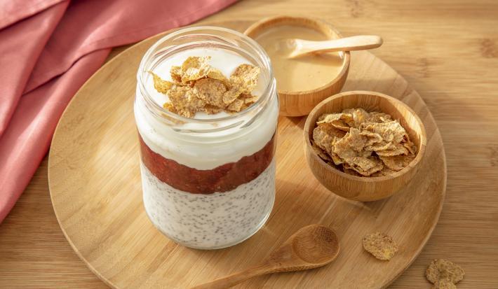 Foto da receita de overnight nesfit cereal. Observa-se um pote com 3 camadas: uma com a chia e iogurte, outra com geleia de frutas vermelhas e outra de iogurte, com NESFIT cereal por cima. Pasta de amendoim e um potinho com o cereal decoram a foto.