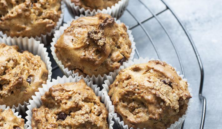 Fotografia de sete muffins com chocolate branco e preto em uma forminha de papel branca, que estão em cima de uma grade redonda na cor preta. Os bolinhos estão sobre uma mesa de cor clara.