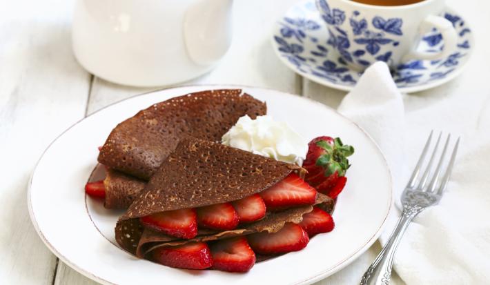 Fotografia de panquecas de chocolate com morango, e um pouco de chantilly ao lado, dentro de um prato branco raso. Ao redor , um garfo sobre uma toalha branca, um bule branco e uma xícara de chá em cima do prato pequeno, ambos são azuis e brancos.