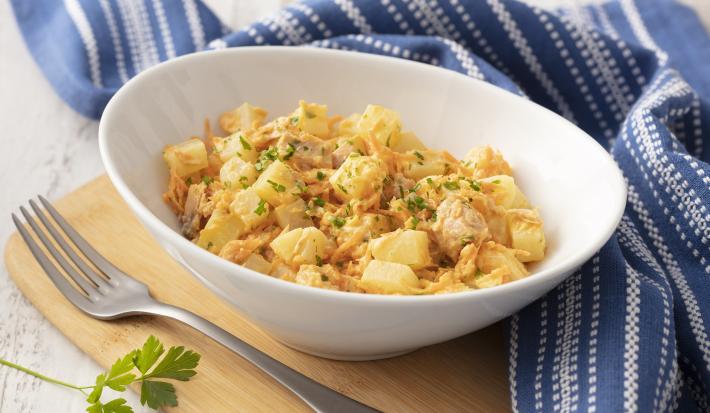 Fotografia em tons de azul em uma bancada de madeira com um pano listrado azul ao lado. No centro, um prato fundo branco com a salada de batata, atum, cebola e salsinha.