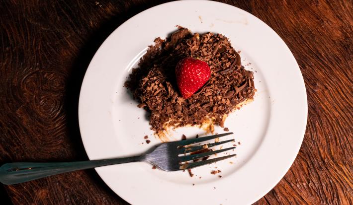 Fotografia vista de cima de um pedaço de pavê com mousse e chocotrio. Por cima tem ganache, raspas de chocolate e morango. O pavê está sobre um prato branco de vidro com um garfo.