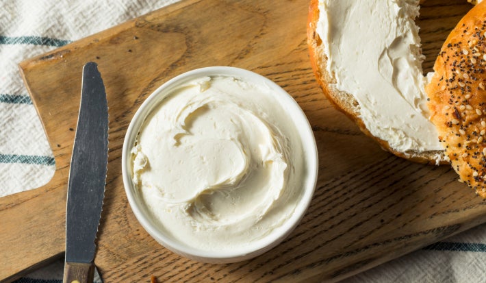 Fotografia mostra um requeijão servio em um pote branco, do seu lado esquerdo há uma faca e do lado direito um bagel com requeijão.