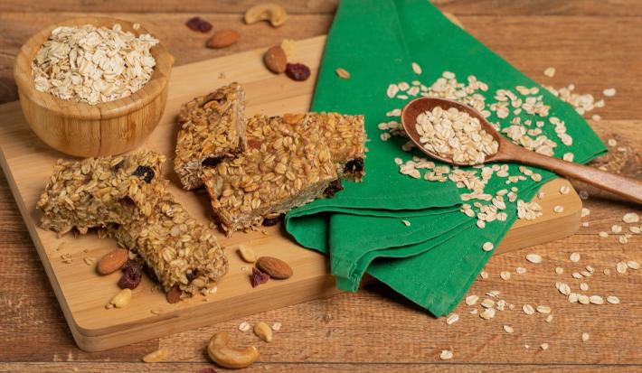 Foto de uma bancada de madeira. Sobre ela há uma tábua de madeira com algumas castanhas espalhadas, flocos de aveia, um tecido verde e a receita de barrinhas de cereal. Ao fundo há um potinho de madeira com mais aveias dentro