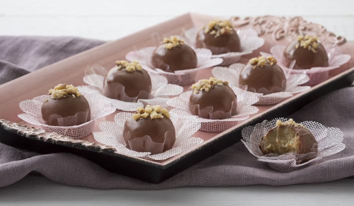 Fotografia em tons de lilás e chocolate, com bandeja lilás claro ao centro contendo bombons de chocolate sobre forminhas rendadas brancas e decorados com nozes picadas, ao lado bombom mordido, sobre guardanapo lilás, tudo bancada em tom lilás.