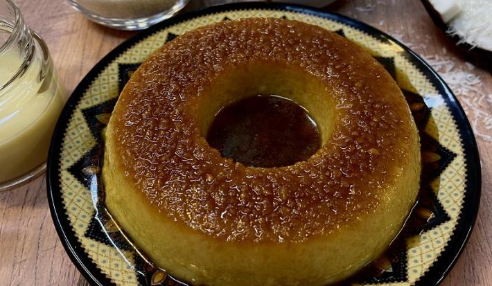 Foto em tons de bege da receita de pudim de tapioca servida em um prato de porcelana amarelo com adornos em marrom. Ao fundo, uma xícara de tapioca granulada, um coco cortado ao meio e um vidro com leite MOÇA