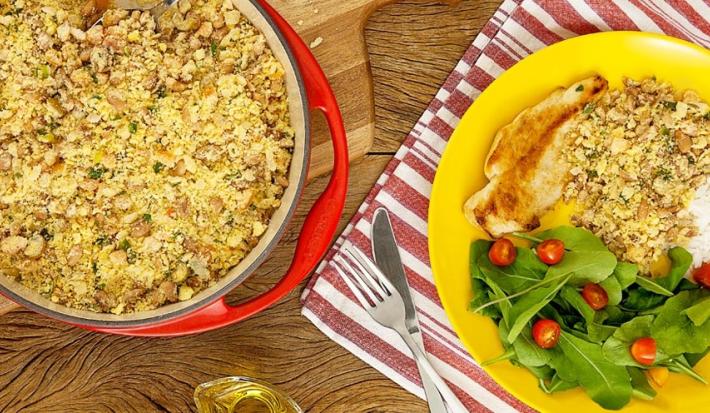 Fotografia em tons de amarelo e vermelho em uma bancada de madeira com um pano vermelho listrado com um prato amarelo com salada verde, arroz, frango e farofa de feijão. Ao lado, uma frigideira vermelha com farofa dentro.