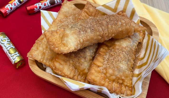 Fotografia em tons de vermelho com um recipiente com vários pastéis recheados com chocolate Baton ao centro. Os pastéis estão polvilhados com açúcar e canela. Ao fundo existe embalagens de chocolate Baton.