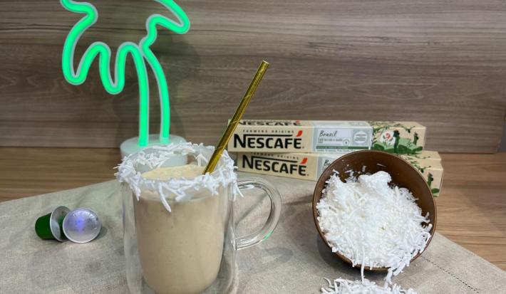 Fotografia em tons de verde com uma caneca de vidro ao centro. Dentro da caneca existe uma mistura de café gelado com leite de coco. Ao lado existe um pote marrom cheio de coco ralado e ao fundo duas caixas de cápsulas NESCAFÉ