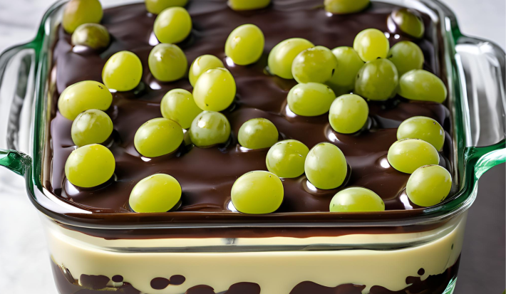 A fotografia mostra um refratário com camadas de uvas verdes sem sementes, cobertas por um creme branco espesso e finalizadas com uma ganache de chocolate ao leite brilhante.