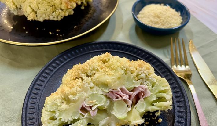 Foto em tons de amarelo claro da receita de couve-flor recheada servida em um prato de porcelana azul escuro com talheres prateados ao lado e um recipiente azul com queijo parmesão ralado. Ao fundo há um prato preto com outra porção da couve-flor