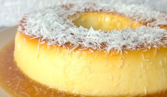 Fotografia em tons claros com um pudim de coco ao centro. O pudim ter cor caramelada e é coberto com coco ralado por cima.