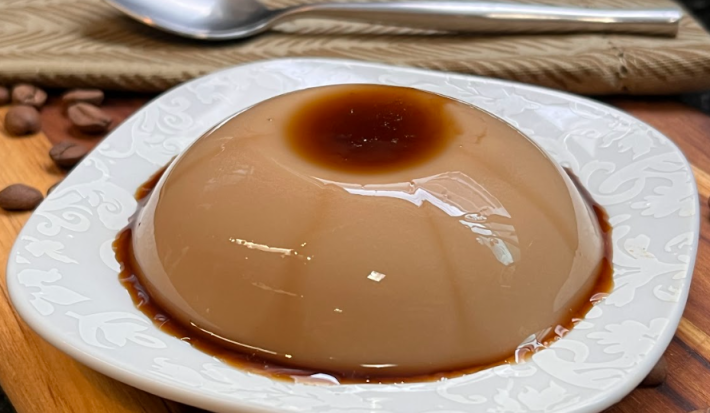 Imagem mostra um pudim redondo e pequeno, coberto de caramelo, em tons castanhos e marrons sobre um prato branco. O prato está apoiado em cima de uma madeira, com grãos de café espalhados, um guardanapo de pano bege e uma colher.