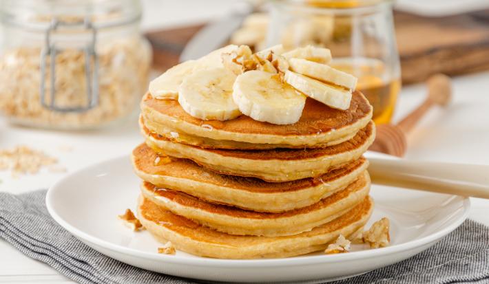 Foto da receita de Panqueca de Banana. Observa-se uma torre de panquecas altas e fofas com banana em cima e um pouco de mel escorrendo.
