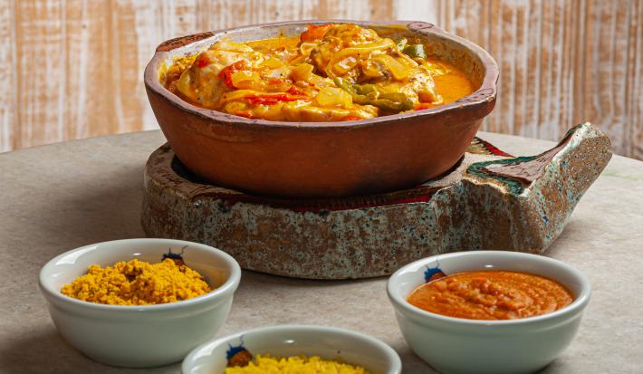 Fotografia de um cozido de legumes e carne dentro de um refratário de cerâmica sobre um apoio feito de pedra. Na frente do cozido, três potes pequenos e brancos de vidro com arroz, farofa e pirão. A comida está sobre uma mesa redonda de cor cinza.