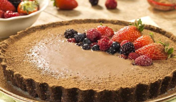Fotografia em tons de marrom em uma mesa com uma toalha bege, um prato raso redondo grande com a torta de biscoito Nesfit com cacau e frutas vermelhas para decorar a torta, em cima do prato.