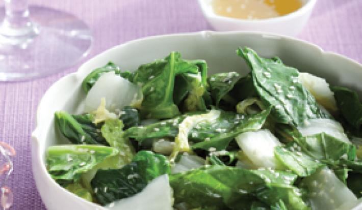 Fotografia em tons de branco, verde e lilás, ao centro uma saladeira com folhas verdes e gergelim, ao lado potinho de azeite, copos e uma jarra, sobre uma toalha lilás,