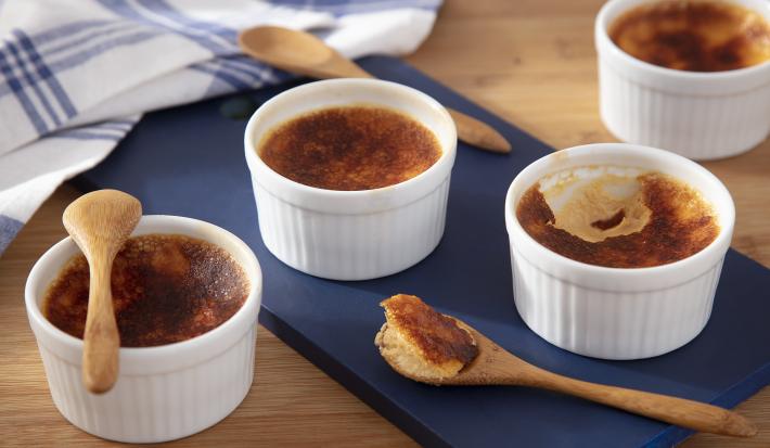 Fotografia em tons de amarelo em uma bancada de madeira de cor marrom. Ao centro, uma tábua de madeira azul contendo o creme brulee. Ao lado, há algumas colheres de pau e ao fundo, um pano azul quadriculado.