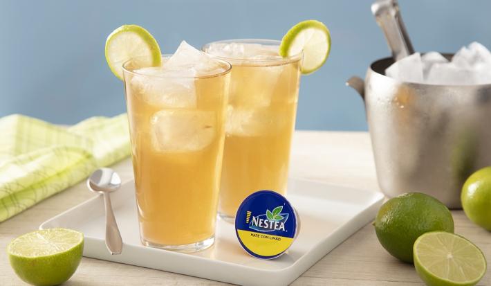 Foto da receita do drink de dolce gusto nesta limão servida em dois copos altos de vidro sobre uma base de porcelana branca com uma colher prateada e uma cápsula de nestea. Ao fundo há um balde cromado de gelo e limões cortados, além de um pano verde.