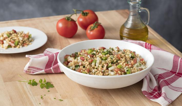 Fotografia em tons de branco e vermelho, ao centro recipiente branco com salada de feijão e tomate, ladeada por prato com salada, três tomates, galheteiro com azeite e guardanapo branco com listras vermelhas, tudo sobre bancada de madeira e fundo cinza.