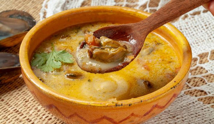 Foto da receita de Sururu ao Leite de Coco. Observa-se uma tigela amarela com a receita bem cremosa e uma colher de pau está mostrando o sururu