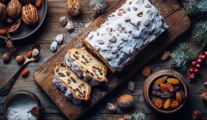 Fotografia de um bolo com frutas secas em uma tábua de madeira, polvilhado com açúcar de confeiteiro. Ao redor, nozes, amêndoas e frutas secas criam um ambiente festivo e acolhedor.