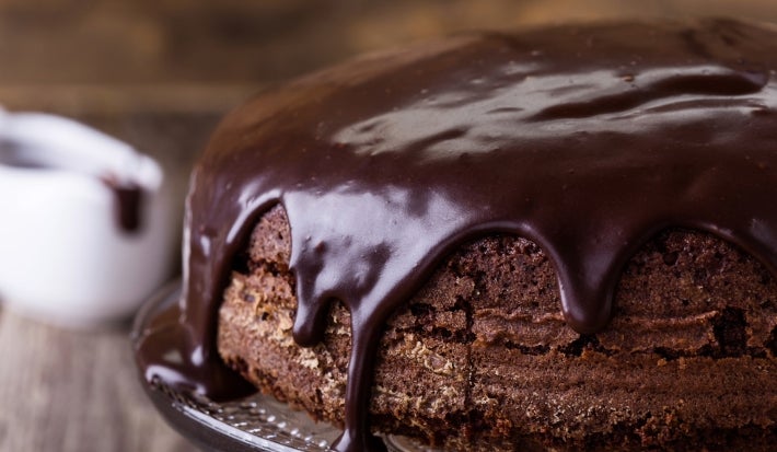 Um bolo de chocolate redondo, com textura densa e caseira, está coberto por uma espessa camada de cobertura de chocolate brilhante que escorre pelas laterais. Ele está sobre um suporte de vidro, pronto para ser servido.