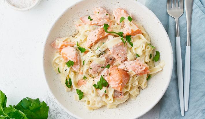 Fotografia em tons de branco, rosa, amarelo e verde de uma bancada branca vista de cima. Ao centro contém um prato redondo com macarrão, pedaços de salmão e salsa.