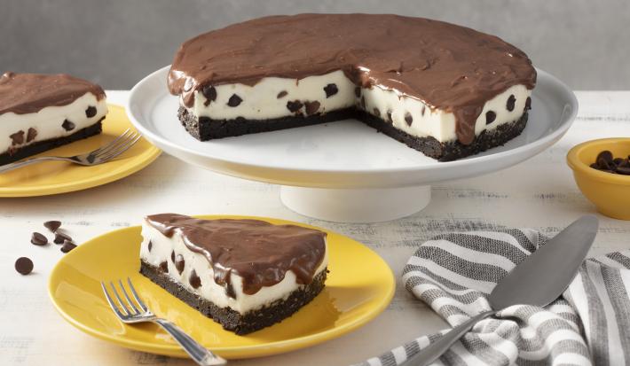 Fotografia em tons de amarelo em uma bancada cinza com um pano listrado em cinza e branco. Ao centro, um prato amarelo com uma fatia da torta de ninho com gotas de chocolate. Atrás um suporte branco com a torta inteira.
