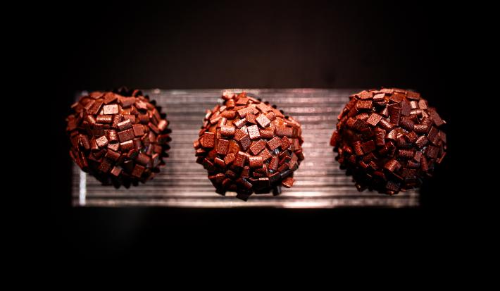 Fotografia vista de cima de três brigadeiros de tucupi reduzido recheados com cupuaçu e enrolados no chocolate amargo. Os brigadeiros estão em um apoio na cor prata, com o fundo escuro.