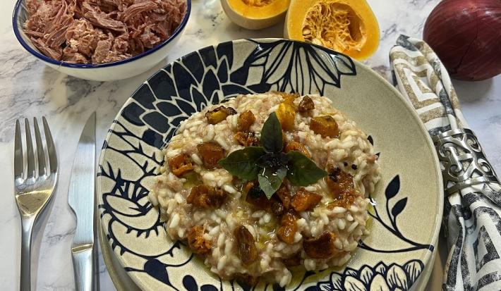 Foto da receita de risoto de carne seca e abóbora em tons de laranja servida em um prato de porcelana azul e branco. Ao lado, há talheres prateados e um guardanapo adornado, além de carne seca, abóbora e cebola ao fundo para decorar.