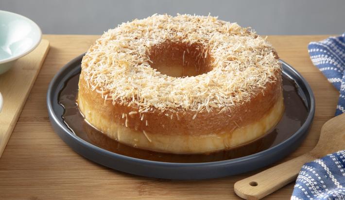 Foto da receita de pudim de tapioca com calda de coco queimado servido em uma porção grande sobre um prato azul escuro em cima de uma mesa de madeira com um paninho azul listrado