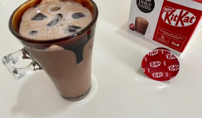 Fotografia em tons de vermelho com uma caneca de vidro ao centro. Dentro da caneca existe uma receita de Iced Mocha feita com Dolce Gusto Kitkat. Nas bordas da caneca existe uma calda de chocolate